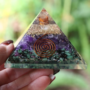 May include: A layered pyramid-shaped object with a clear base. The layers include green, purple, and gold-colored materials, with a copper coil at the center. The pyramid is held in a hand against a blurred green background.