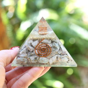 May include: A clear resin pyramid with a copper spiral and coil design. The pyramid is filled with small, light-colored stones and gold flakes. The pyramid is held in a hand against a blurred green background.