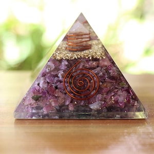 May include: A clear pyramid-shaped resin sculpture filled with crushed purple stones and gold flakes. A copper spiral and a small crystal are embedded within the pyramid. The sculpture is displayed on a wooden surface.