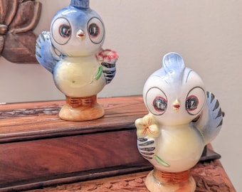 May include: Two ceramic bird figurines. Each bird has a blue head and wings, a yellow body, and a brown base. They hold a flower in their wing. The figurines are on a wooden surface.