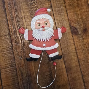 May include: A red and white pull-string Santa Claus toy with a white beard and black shoes. The toy is on a wooden surface.