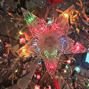 May include: A clear, multi-faceted star tree topper with a warm, colorful glow. The star is surrounded by silver tinsel, string lights, and other festive ornaments. The overall theme is Christmas.
