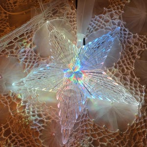 May include: A clear, faceted star-shaped Christmas tree topper with internal rainbow-colored lights. The star has a pointed top and five radiating points. A white plastic base and black cord are attached.