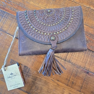 May include: A brown leather wallet with a decorative embossed design and a tassel. The wallet has a flap closure and is adorned with metal studs. The brand name "Patricia Nash" is visible on a tag attached to the wallet.