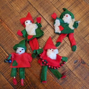 May include: Four vintage Christmas elf ornaments. Each elf has a red hat, green jacket, and red pants. They have white beards and are decorated with beads and hearts. The ornaments are on a wooden surface.