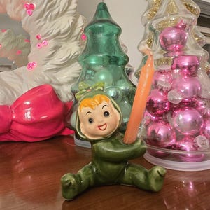 May include: A vintage ceramic figurine of a seated child in a green outfit, holding an orange candle. The figurine has a smiling face and is surrounded by Christmas tree decorations, including glass trees and pink ornaments.