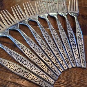 May include: A collection of stainless steel forks with a decorative etched design on the handles. The forks are arranged on a wooden surface, showcasing their polished finish and intricate detailing. The tines are evenly spaced.