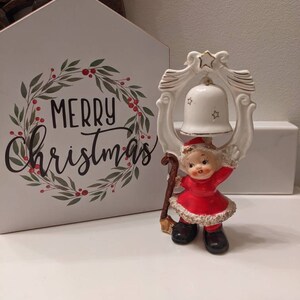 May include: A vintage Christmas figurine of a child in a red dress and black boots holding a white bell with gold accents. The figurine is set against a white house-shaped sign with the words "MERRY Christmas" in black script.