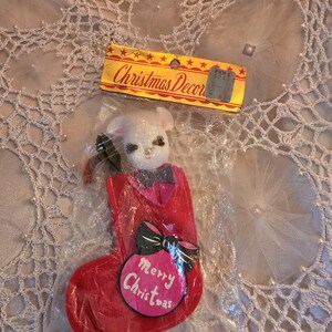 May include: A vintage Christmas stocking ornament in a plastic wrapper. The red stocking has a pink ornament with "Merry Christmas" text and a black bow. A white bunny head with a black bow tie is at the top. The tag reads "Christmas Decor".