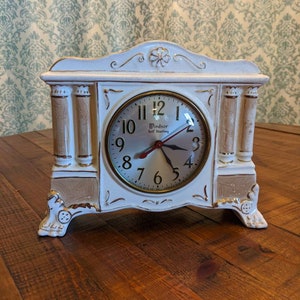 May include: White ceramic clock with gold accents and a white face. The clock has a gold rim and says "Windsor Self Starting" on the face.