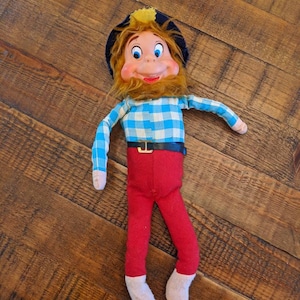 May include: A vintage doll with a painted face, orange hair, and a yellow tuft on a blue hat. The doll wears a blue and white plaid shirt, red pants, a black belt, and white socks. The doll is approximately 12 inches tall.