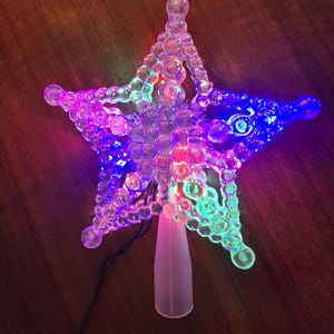May include: A clear, multi-colored star-shaped Christmas tree topper with a conical base. The star is illuminated with red, blue, green, and purple lights. The topper is made of clear plastic and has a series of bubble-like shapes.