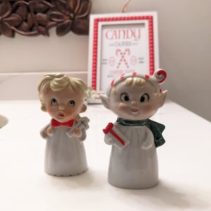 May include: Two vintage porcelain figurines of Christmas elves. One elf is wearing a white robe with a red bow tie and holding a red and gold gift. The other elf is wearing a white robe with a green scarf and a red and white striped hat and holding a red and white striped gift. The sign behind the elves reads "Candy Canes".