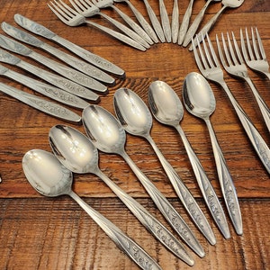 May include: A collection of stainless steel silverware, including forks, knives, and spoons, arranged on a wooden surface. The silverware features a floral design on the handles. The spoons have a reflective, polished surface.