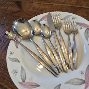 May include: A set of vintage silverware, including spoons, forks, and a knife, arranged on a white plate with a pink and gray leaf pattern. The silverware has a starburst design on the handles, and the plate is approximately 10 inches in diameter.