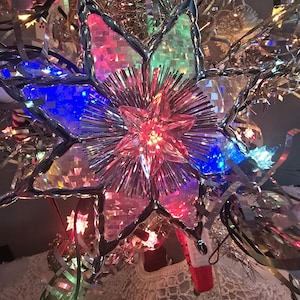 May include: A star-shaped Christmas tree topper with a silver frame and colorful lights. The topper has a central red starburst and is surrounded by tinsel and other decorative elements. The lights are red, blue, and green.