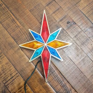 May include: A colorful star-shaped Christmas tree topper with red, blue, and yellow segments. The star has a silver trim and a black electrical cord. The topper is placed on a wooden surface.