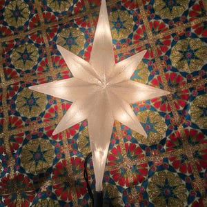 May include: A glowing, frosted glass star tree topper with eight points. The star is lit from within and sits atop a black base with a clip. The background is a patterned fabric with red, blue, and gold geometric designs.