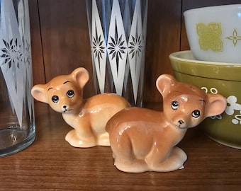 May include: Two ceramic lion cub figurines with glossy brown fur and black eyes. The cubs are in a seated position, with their ears perked up. The figurines are displayed on a wooden shelf, alongside decorative glassware and bowls.