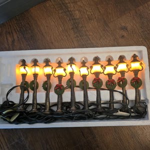 May include: A string of ten miniature decorative street lamps connected by a black electrical cord. Each lamp features a bronze-colored post, a glowing yellow light, and a small green wreath adorned with a red bow at the base of the lantern.