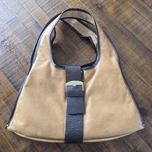 May include: Tan leather shoulder bag with a dark brown strap and trim. The bag features a rectangular dark brown strap with a gold-colored buckle. The bag has a triangular shape and a long strap for carrying over the shoulder.