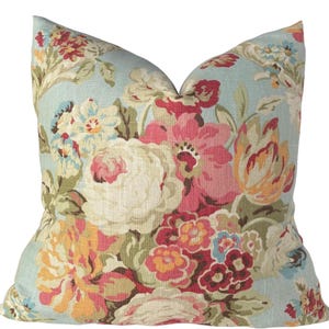 May include: A square throw pillow featuring a light blue background with a dense, colorful floral pattern. The design includes large, blooming flowers in shades of pink, cream, yellow, and deep red, accented by green leaves and stems.
