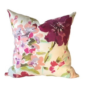 May include: A square decorative pillow with a floral pattern. The pillow features a cream-colored background with watercolor-style flowers in shades of pink, purple, and burgundy. Green leaves and stems are also visible.