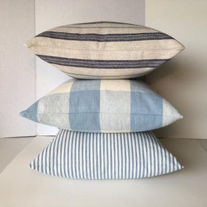 May include: Three decorative throw pillows stacked on top of each other. The top pillow is a cream and navy blue striped pillow. The middle pillow is a blue and white plaid pillow. The bottom pillow is a white and blue striped pillow.