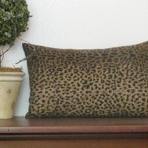 May include: A rectangular decorative pillow with a leopard print pattern in shades of brown and black. The pillow is placed on a dark wooden surface, with a small potted plant to the left.