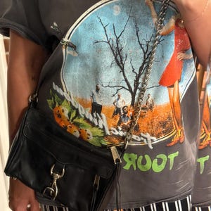 May include: A black leather crossbody bag featuring a silver-tone chain strap, a front flap with a metallic clasp, and a side zipper detail. The bag is worn over a dark gray t-shirt with a graphic print of a tree, figures, and orange fruit.
