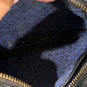 May include: The interior of a black bag featuring a blue fabric lining with a black polka dot pattern. The bag has a gold-toned metal zipper closure along the top edge, which is partially visible. The interior space is open and shows a seam.