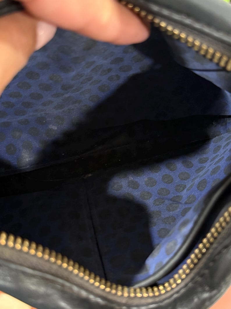 May include: The interior of a black bag featuring a blue fabric lining with a black polka dot pattern. The bag has a gold-toned metal zipper closure along the top edge, which is partially visible. The interior space is open and shows a seam.