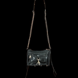 May include: A black leather crossbody bag featuring a gold-tone chain strap, a front zippered pocket, and a central metal clasp closure. The bag includes long leather zipper pulls and gold-tone hardware accents, suspended against a black background.