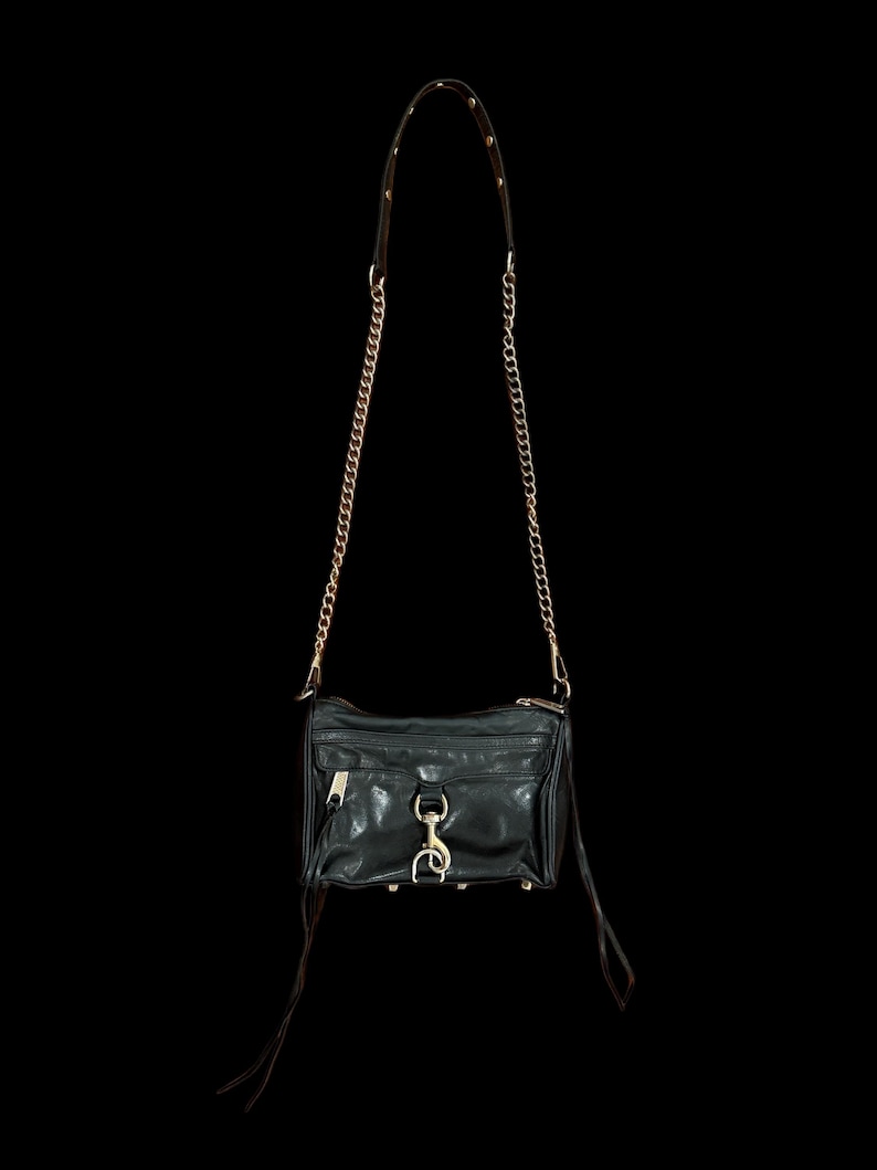 May include: A black leather crossbody bag featuring a gold-tone chain strap, a front zippered pocket, and a central metal clasp closure. The bag includes long leather zipper pulls and gold-tone hardware accents, suspended against a black background.