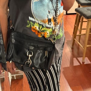 May include: A black leather crossbody bag featuring a silver-toned metal chain strap, a front flap with a prominent silver lobster clasp closure, and a side zipper detail with long leather pulls. The bag has a structured rectangular shape.