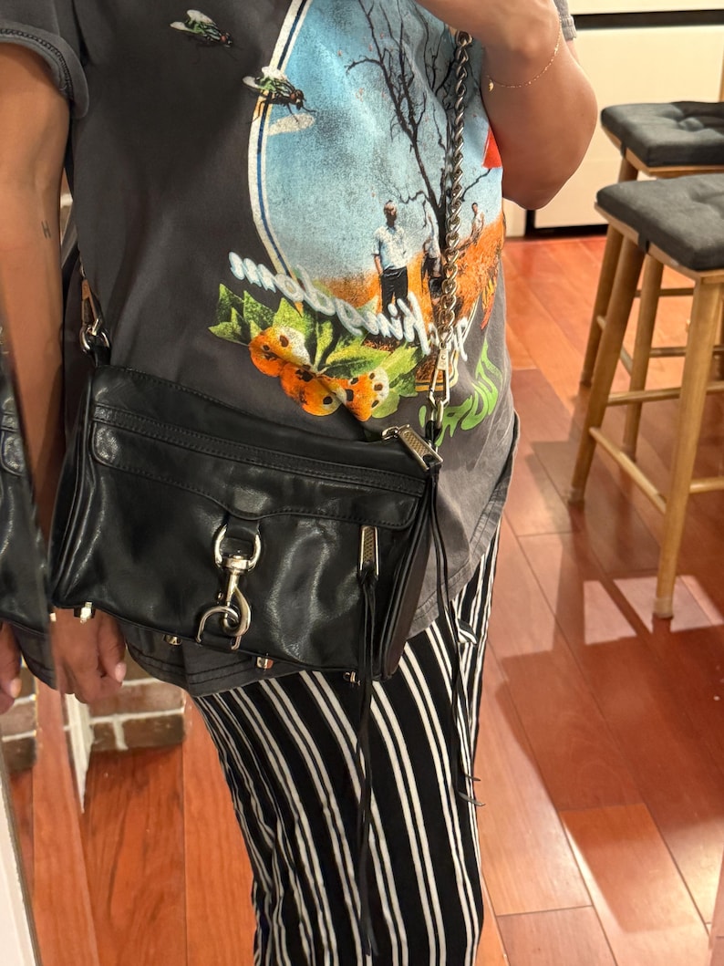 May include: A black leather crossbody bag featuring a silver-toned metal chain strap, a front flap with a prominent silver lobster clasp closure, and a side zipper detail with long leather pulls. The bag has a structured rectangular shape.
