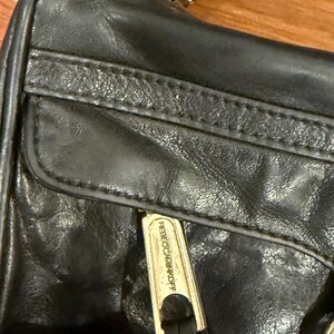 May include: A black leather handbag featuring a front zippered pocket with a metallic pull tab engraved with the text REBECCAMINKOFF. The bag includes a top zipper closure with a matching metallic pull tab also displaying the brand name.