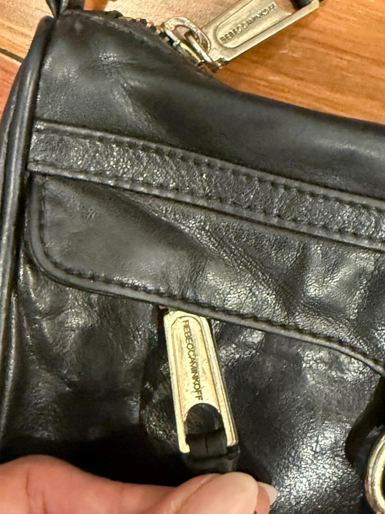 May include: A black leather handbag featuring a front zippered pocket with a metallic pull tab engraved with the text REBECCAMINKOFF. The bag includes a top zipper closure with a matching metallic pull tab also displaying the brand name.