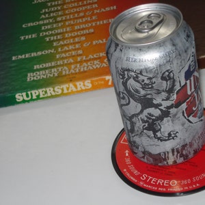 Puede incluir: Una lata de cerveza plateada con un gr&aacute;fico de le&oacute;n en blanco y negro y el texto "Beer Before Glory". La lata est&aacute; sobre un disco de vinilo rojo con el texto "360 Sound Stereo 360 Sound" y "Columbia Marcas Reg. Printed in U.S.A."