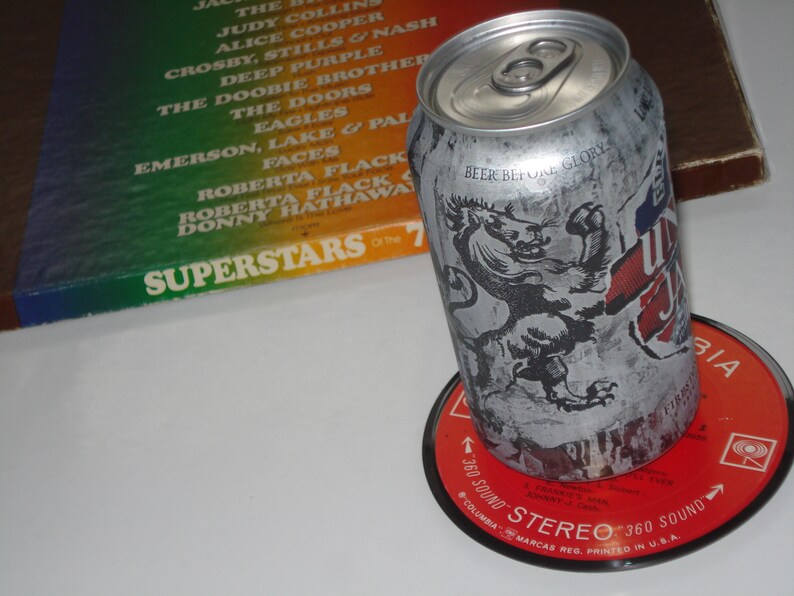 Puede incluir: Una lata de cerveza plateada con un gr&aacute;fico de le&oacute;n en blanco y negro y el texto "Beer Before Glory". La lata est&aacute; sobre un disco de vinilo rojo con el texto "360 Sound Stereo 360 Sound" y "Columbia Marcas Reg. Printed in U.S.A."