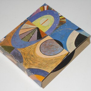May include: A cardboard box with a colorful abstract design. The artwork features a mix of blue, orange, yellow, and purple shapes. The box is angled, showing the patterned top and the plain brown sides.