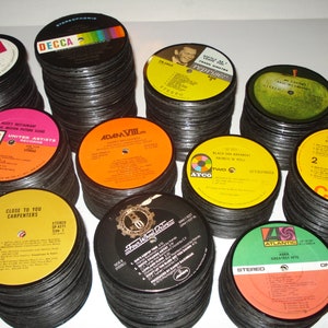 Puede incluir: Una colecci&oacute;n de posavasos de vinilo vintage. Los posavasos est&aacute;n apilados en pilas y presentan varias portadas de &aacute;lbumes, incluyendo "Hurrah!" de The Hollies, "Adam VIII" de Adam Faith, "Close to You" de The Carpenters, "The Beach Boys Christmas Album" de The Beach Boys y "Greatest Hits" de ABBA.