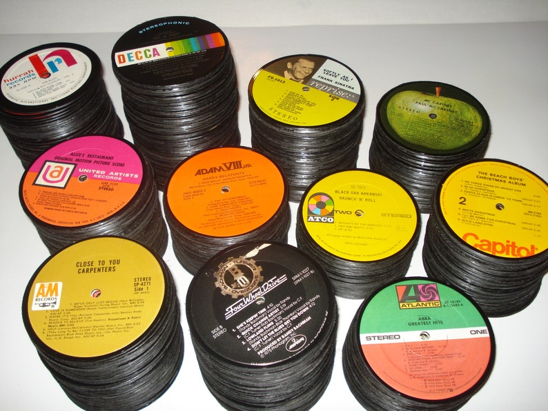 Puede incluir: Una colecci&oacute;n de posavasos de vinilo vintage. Los posavasos est&aacute;n apilados en pilas y presentan varias portadas de &aacute;lbumes, incluyendo "Hurrah!" de The Hollies, "Adam VIII" de Adam Faith, "Close to You" de The Carpenters, "The Beach Boys Christmas Album" de The Beach Boys y "Greatest Hits" de ABBA.