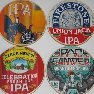 May include: Four round beer coasters with different designs. One coaster features "IPA" with a person on a creature. Another says "Firestone Union Jack IPA". A third says "Sierra Nevada Celebration IPA". The last says "Space Camper Quantum Hop IPA".