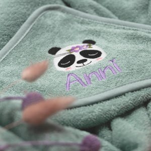 Nahaufnahme der Kapuze von einem salbeifarbenen Handtuch. Als Motiv wurde ein Pandabär mit fliederfarbenem Blumenschmuck gestickt. Der Name "Anni" wurde ebenfalls in flieder in einer hübschen Druckschrift gestickt.