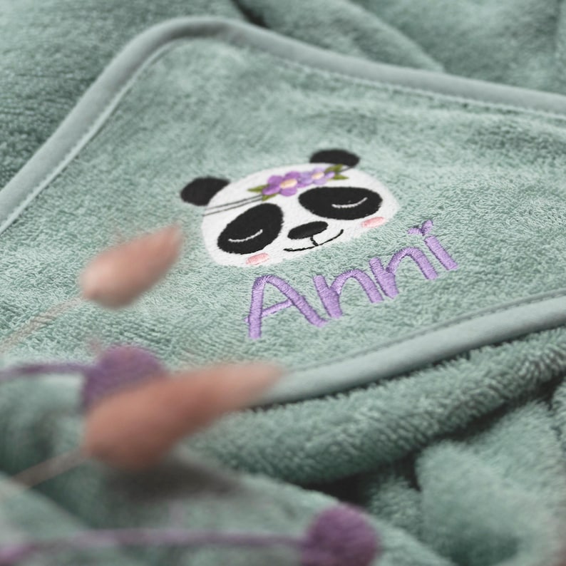 Nahaufnahme der Kapuze von einem salbeifarbenen Handtuch. Als Motiv wurde ein Pandabär mit fliederfarbenem Blumenschmuck gestickt. Der Name "Anni" wurde ebenfalls in flieder in einer hübschen Druckschrift gestickt.