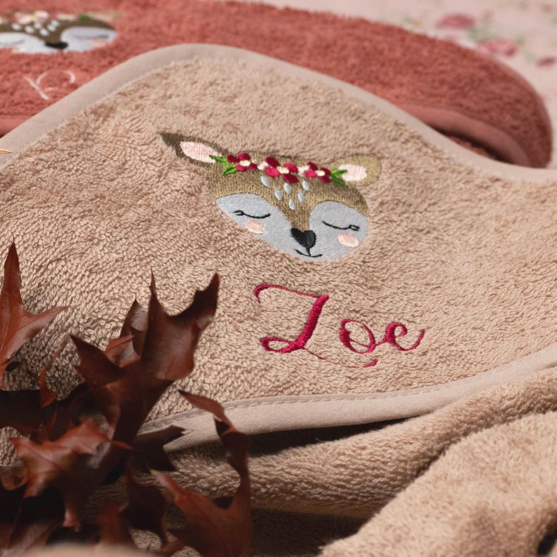 Nahaufnahme der Kapuze von einem rosenholzfarbenen Handtuch. Als Motiv wurde ein Reh mit rotem Blumenstirnband gestickt. Der Name "Zoe" wurde ebenfalls in rot in einer hübschen Schreibschrift gestickt.
