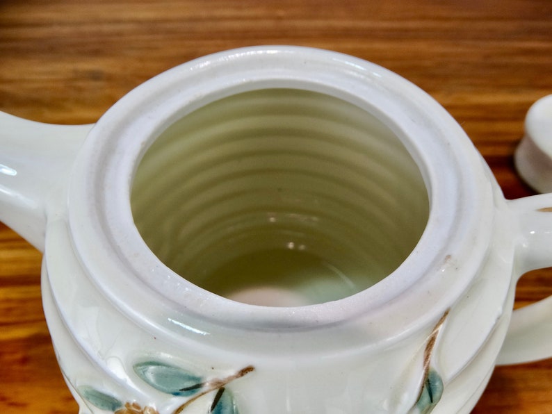 May include: White ceramic teapot with a floral design. The teapot has a wide opening and a handle on the side.