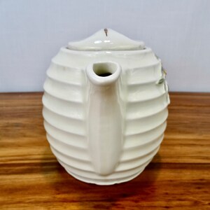 May include: White ceramic teapot with a ribbed design and a floral pattern. The teapot has a spout and a lid.