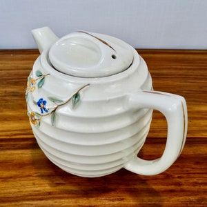 May include: White ceramic teapot with a ribbed design and floral accents. The teapot has a lid with a small hole in the top and a gold-trimmed handle.